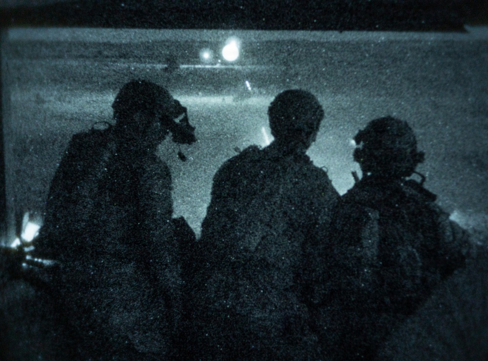 A night-vision or low-light photograph showing three silhouetted figures in military gear observing what appears to be a helicopter in the background. The image has a characteristic blue-green tint and heavy grain consistent with night-vision or infrared photography. The figures appear to be equipped with helmets, communications gear, and tactical equipment.