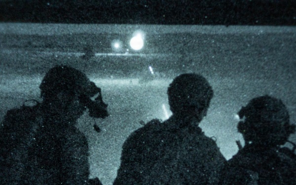 A night-vision or low-light photograph showing three silhouetted figures in military gear observing what appears to be a helicopter in the background. The image has a characteristic blue-green tint and heavy grain consistent with night-vision or infrared photography. The figures appear to be equipped with helmets, communications gear, and tactical equipment.
