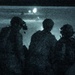 A night-vision or low-light photograph showing three silhouetted figures in military gear observing what appears to be a helicopter in the background. The image has a characteristic blue-green tint and heavy grain consistent with night-vision or infrared photography. The figures appear to be equipped with helmets, communications gear, and tactical equipment.
