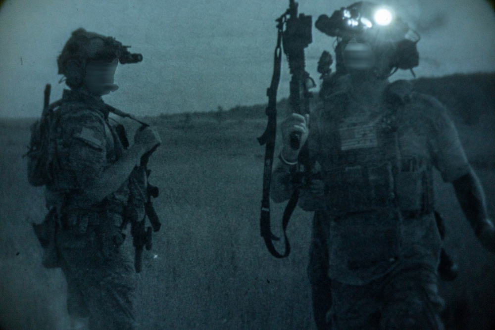 A night-vision style photograph showing two heavily equipped military personnel standing in an open field or terrain. Both individuals are wearing tactical gear including helmets with night-vision devices mounted on them. The image has a characteristic blue-gray monochromatic tone consistent with low-light or night photography.