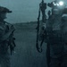 A night-vision style photograph showing two heavily equipped military personnel standing in an open field or terrain. Both individuals are wearing tactical gear including helmets with night-vision devices mounted on them. The image has a characteristic blue-gray monochromatic tone consistent with low-light or night photography.