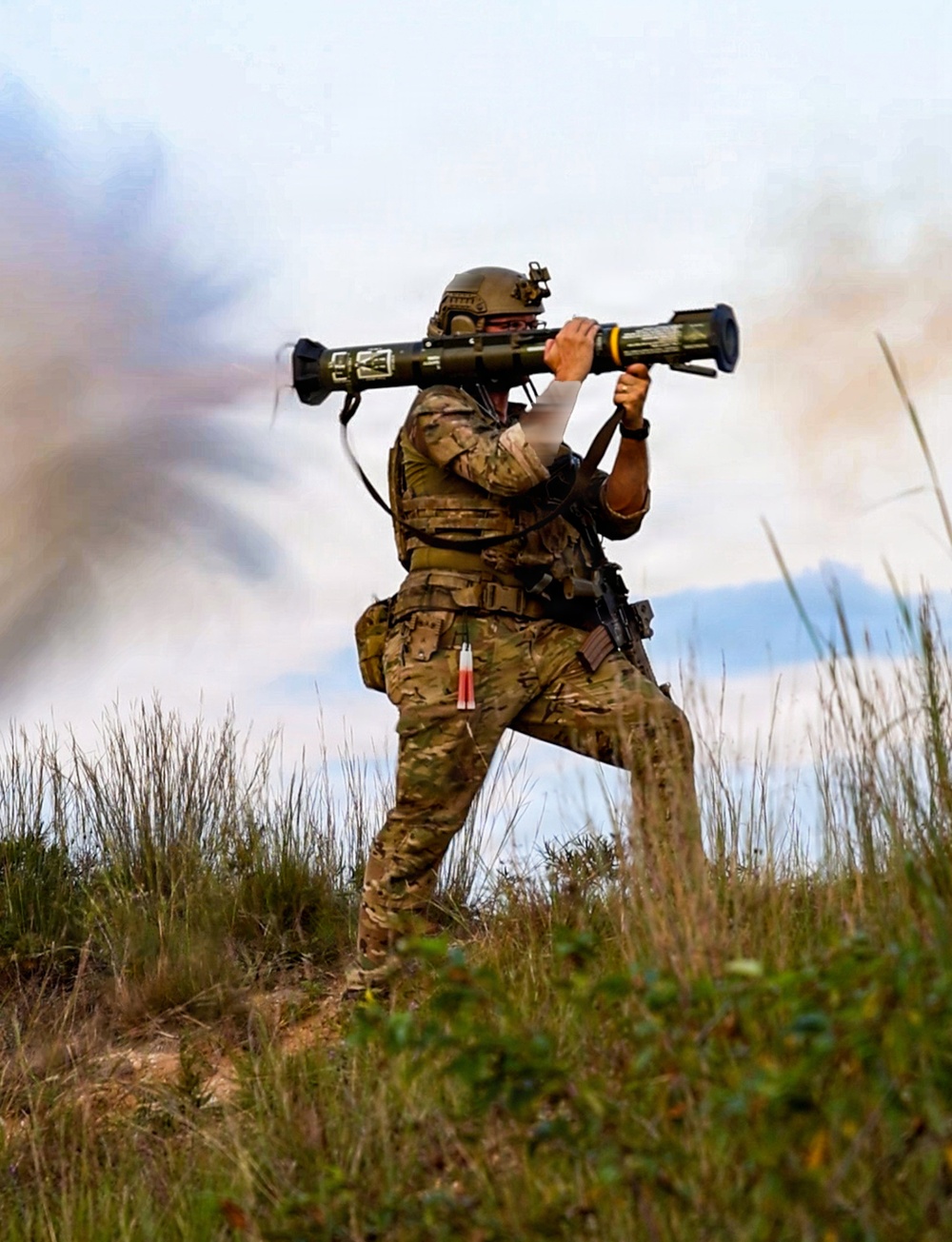 A soldier in full combat gear is firing a shoulder-launched rocket/anti-tank weapon system, with visible backblast smoke and exhaust cloud to the left. The individual is equipped with multicam camouflage, tactical vest, helmet with night-vision mount, and a sidearm. The setting appears to be an outdoor training range or field environment with scrubland vegetation.