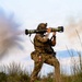 A soldier in full combat gear is firing a shoulder-launched rocket/anti-tank weapon system, with visible backblast smoke and exhaust cloud to the left. The individual is equipped with multicam camouflage, tactical vest, helmet with night-vision mount, and a sidearm. The setting appears to be an outdoor training range or field environment with scrubland vegetation.