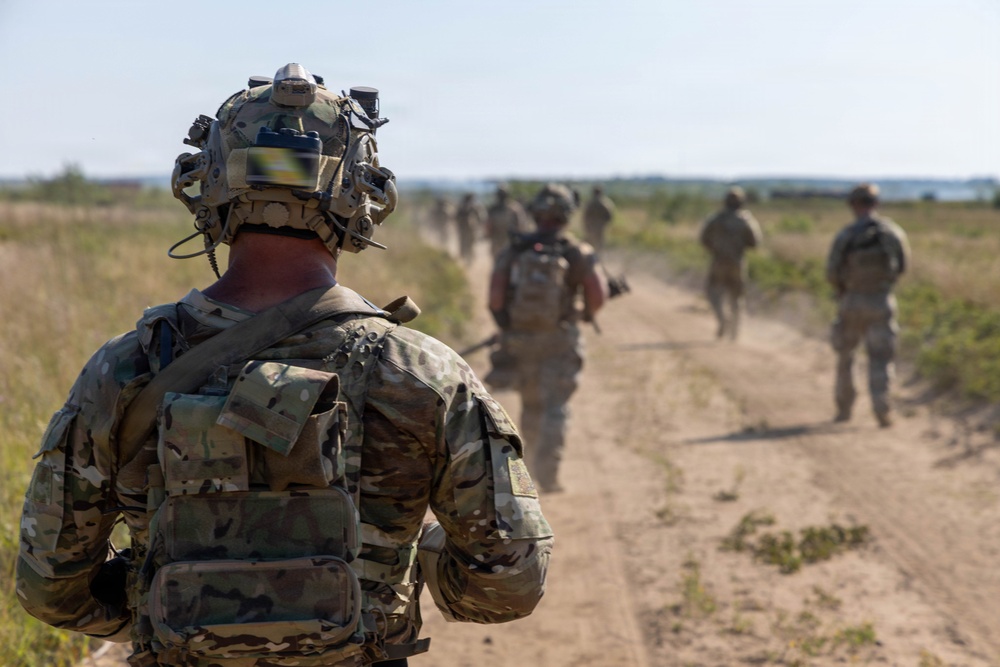 A group of military personnel in full combat gear are conducting a foot patrol along a dusty dirt road through an open field. The foreground soldier is heavily equipped with advanced helmet-mounted devices, body armor, and tactical gear. The column of soldiers extends into the background, suggesting a structured patrol formation.