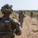 A group of military personnel in full combat gear are conducting a foot patrol along a dusty dirt road through an open field. The foreground soldier is heavily equipped with advanced helmet-mounted devices, body armor, and tactical gear. The column of soldiers extends into the background, suggesting a structured patrol formation.