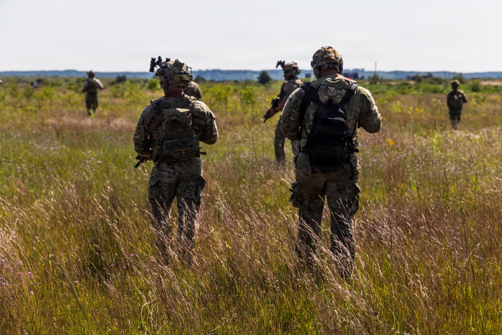 A group of military personnel in full combat gear are conducting a patrol or field exercise through an open grassland area. At least five soldiers are visible moving in a dispersed formation across the terrain. The image appears to depict a training exercise or operational movement in a rural, open environment.