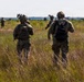 A group of military personnel in full combat gear are conducting a patrol or field exercise through an open grassland area. At least five soldiers are visible moving in a dispersed formation across the terrain. The image appears to depict a training exercise or operational movement in a rural, open environment.