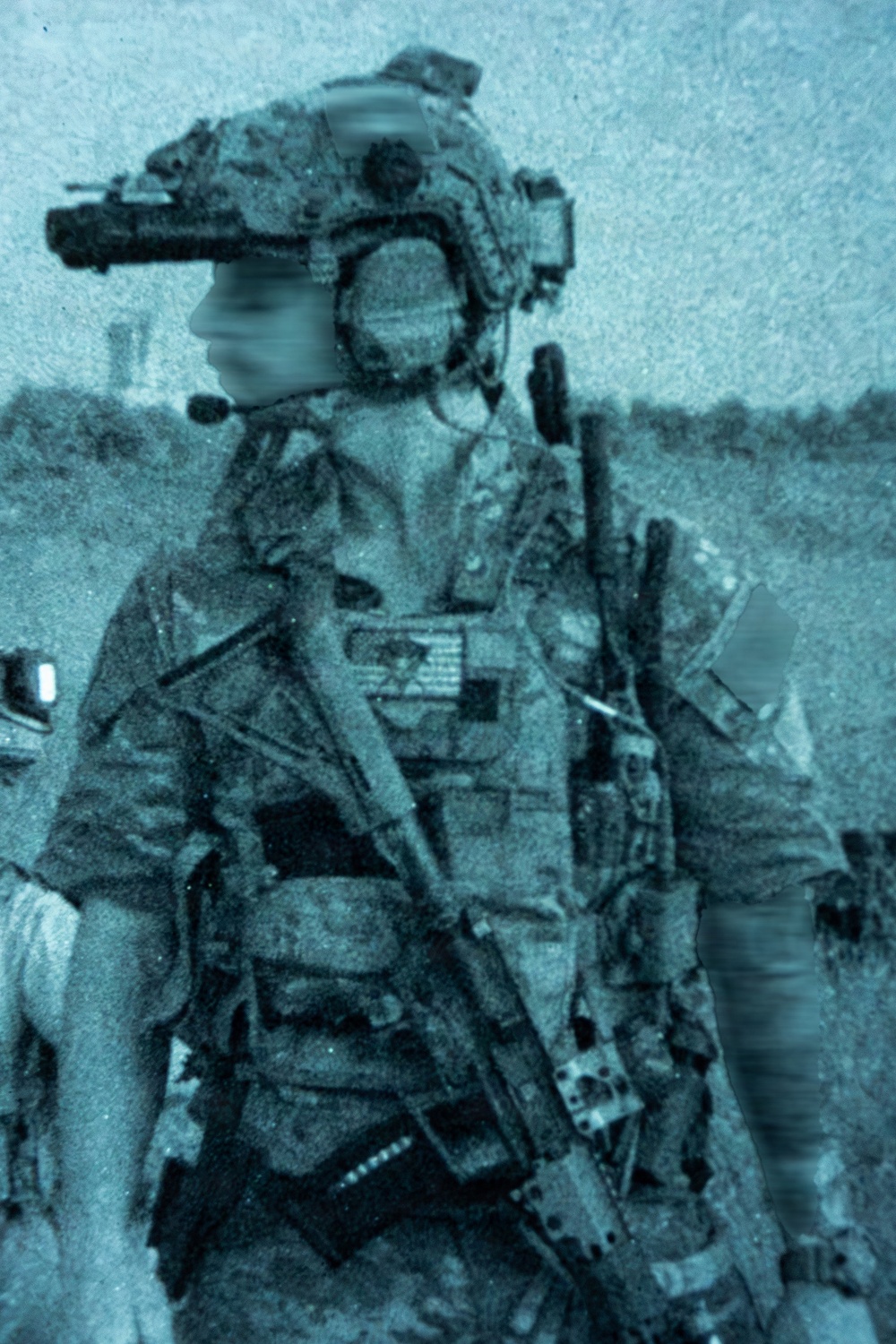 A heavily equipped military operator is depicted in a blue-tinted, grainy photograph with the face deliberately blurred for anonymity. The individual is wearing full tactical gear including a helmet with night-vision device mount, body armor, and carrying a rifle. A second partially visible figure appears in the background to the right.