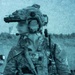 A heavily equipped military operator is depicted in a blue-tinted, grainy photograph with the face deliberately blurred for anonymity. The individual is wearing full tactical gear including a helmet with night-vision device mount, body armor, and carrying a rifle. A second partially visible figure appears in the background to the right.