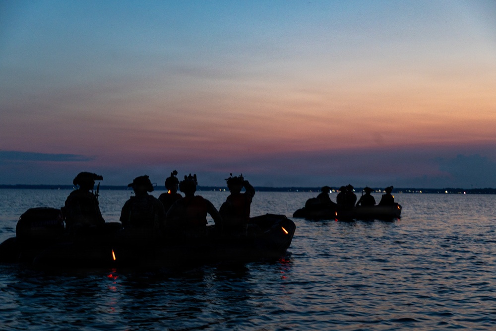 The image shows silhouetted figures in what appear to be two inflatable military-style rubber boats on calm water at dusk or dawn. The personnel are wearing helmets and tactical gear, suggesting a military or special operations maritime exercise. The scene is backlit by a vivid sunset with orange, pink, and blue gradient sky.
