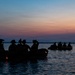 The image shows silhouetted figures in what appear to be two inflatable military-style rubber boats on calm water at dusk or dawn. The personnel are wearing helmets and tactical gear, suggesting a military or special operations maritime exercise. The scene is backlit by a vivid sunset with orange, pink, and blue gradient sky.