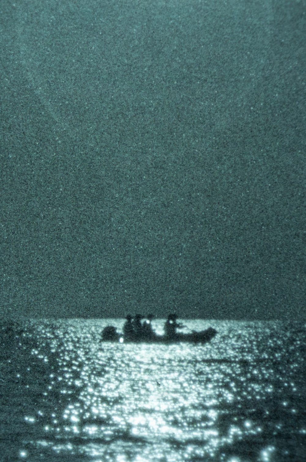 A heavily grainy, teal-toned photograph showing the silhouette of a small boat with multiple occupants on open water. Light reflects off the water's surface in the foreground, creating a bright, sparkling path toward the vessel. The image has a nocturnal or low-light quality, possibly captured with night-vision or infrared equipment.