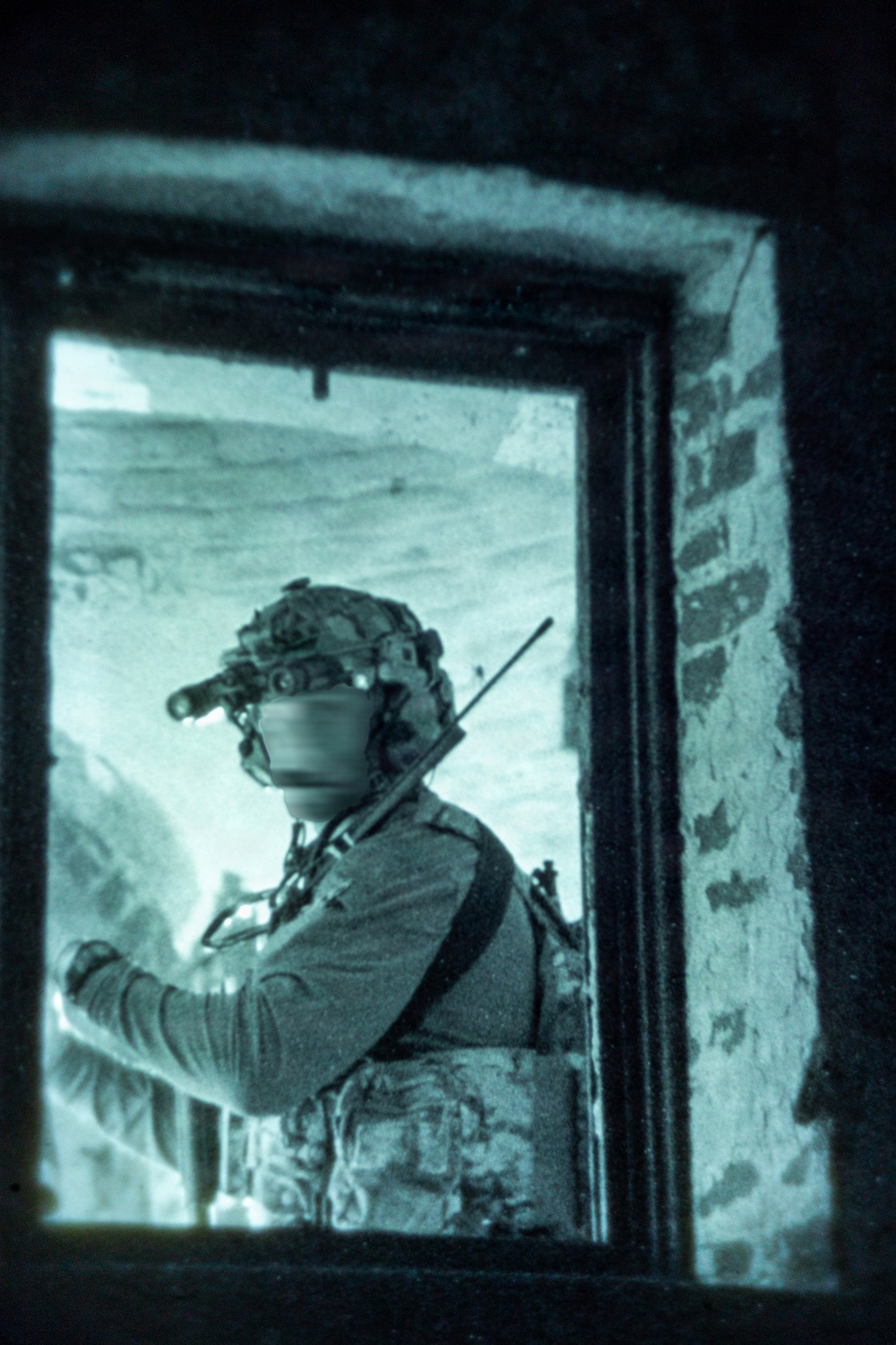 A military or law enforcement operator is visible through a window opening in a stone or brick structure, photographed under night-vision or infrared imaging conditions. The subject is equipped with tactical gear including a helmet-mounted night vision device, body armor, and a slung long-arm weapon. The teal-green monochromatic tone is characteristic of night-vision optics or post-processing to simulate such imagery.