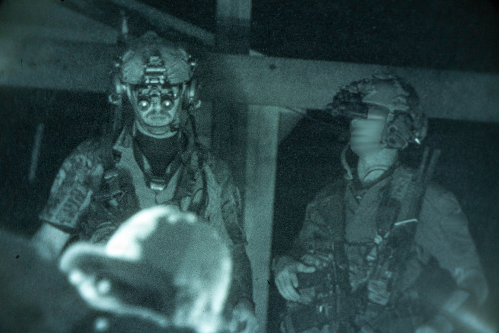 A night-vision captured image showing two military personnel in full tactical gear, including helmets and night-vision devices mounted on their headgear. Both individuals are wearing camouflage uniforms and tactical vests. The image has the characteristic green-tinted, grainy appearance of night-vision photography.