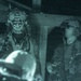 A night-vision captured image showing two military personnel in full tactical gear, including helmets and night-vision devices mounted on their headgear. Both individuals are wearing camouflage uniforms and tactical vests. The image has the characteristic green-tinted, grainy appearance of night-vision photography.