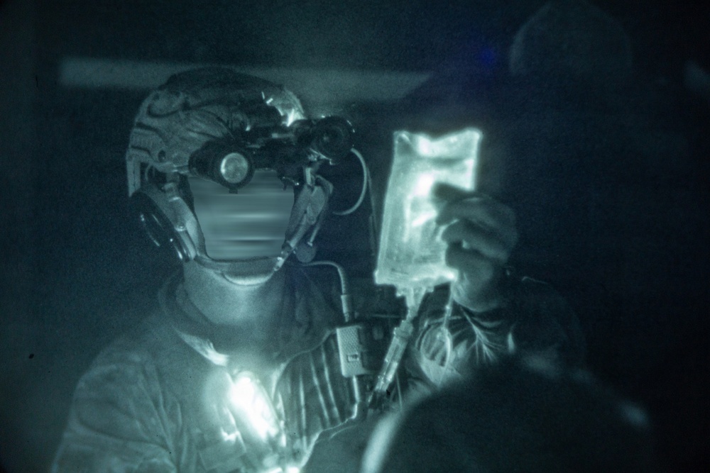A night-vision captured image showing a military medic or combat search and rescue personnel holding up what appears to be an IV fluid bag. The individual is wearing a tactical helmet equipped with night vision goggles (NVGs) and communications headset. The scene suggests an in-flight medical treatment scenario, likely aboard a military helicopter.