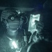 A night-vision captured image showing a military medic or combat search and rescue personnel holding up what appears to be an IV fluid bag. The individual is wearing a tactical helmet equipped with night vision goggles (NVGs) and communications headset. The scene suggests an in-flight medical treatment scenario, likely aboard a military helicopter.