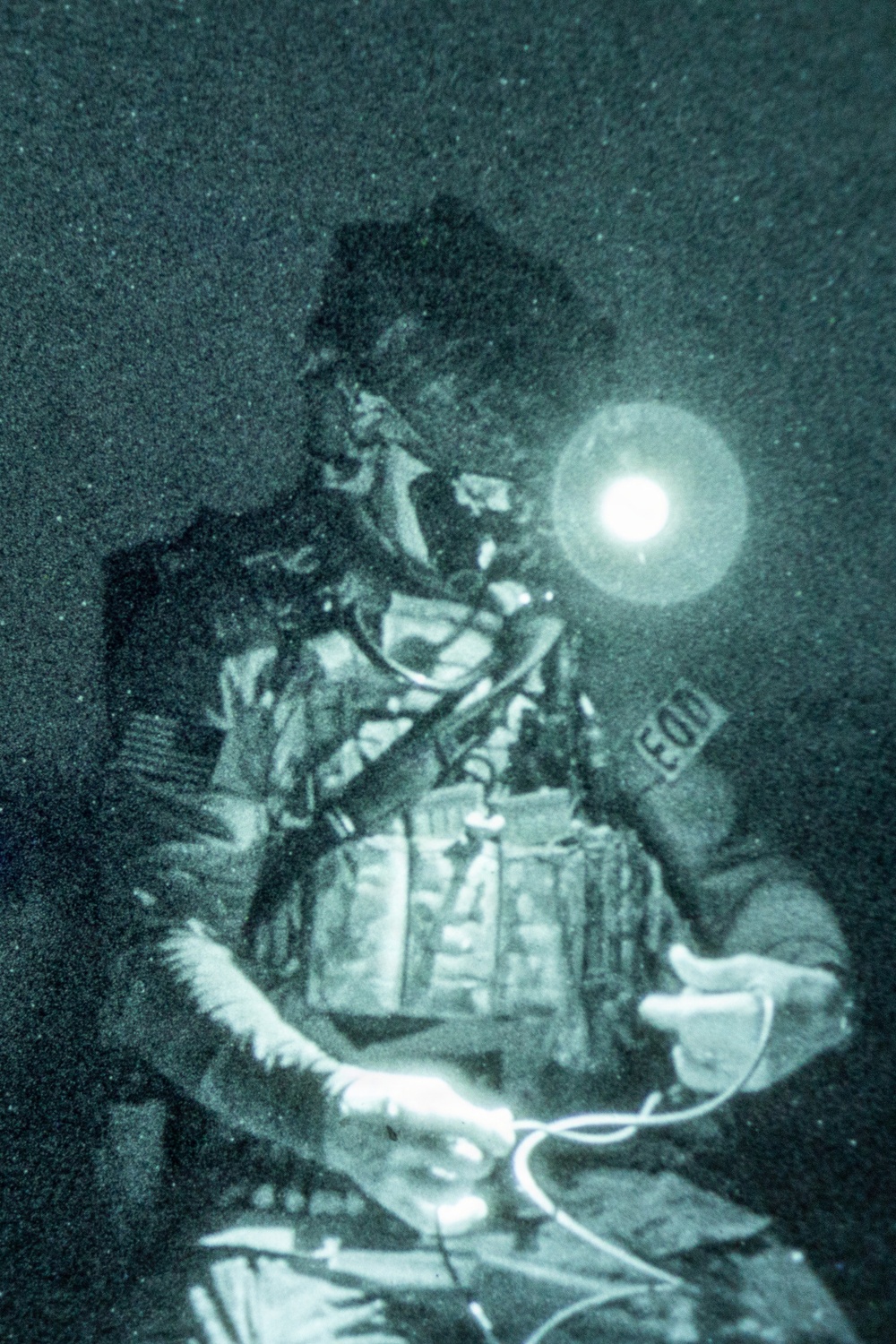 A night-vision or green-tinted photograph showing a military EOD (Explosive Ordnance Disposal) technician working at night. The individual is wearing full tactical gear including a helmet with mounted equipment, body armor with MOLLE pouches, and an EOD patch visible on the arm. The technician appears to be handling wires or cables, consistent with ordnance disposal or demolition work.