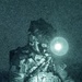 A night-vision or green-tinted photograph showing a military EOD (Explosive Ordnance Disposal) technician working at night. The individual is wearing full tactical gear including a helmet with mounted equipment, body armor with MOLLE pouches, and an EOD patch visible on the arm. The technician appears to be handling wires or cables, consistent with ordnance disposal or demolition work.