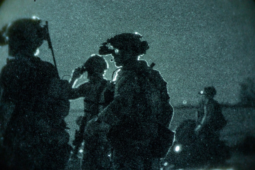 A night-vision captured image showing multiple armed military personnel in full tactical gear operating in darkness. The green-tinted, grainy imagery is characteristic of night-vision optics or image intensifier technology. At least three silhouetted figures are visible, with one prominently displaying night-vision goggles mounted on a helmet.