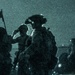 A night-vision captured image showing multiple armed military personnel in full tactical gear operating in darkness. The green-tinted, grainy imagery is characteristic of night-vision optics or image intensifier technology. At least three silhouetted figures are visible, with one prominently displaying night-vision goggles mounted on a helmet.