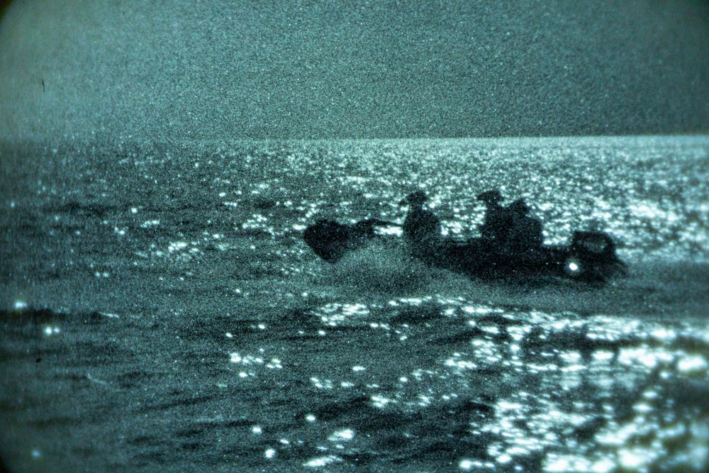 A night-vision or low-light photograph showing a small watercraft on open water. The image captures silhouetted figures aboard what appears to be a small motorized boat or inflatable craft moving across a reflective sea surface. The teal-green tint and heavy grain are consistent with night-vision optics or thermal imaging equipment.