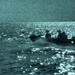 A night-vision or low-light photograph showing a small watercraft on open water. The image captures silhouetted figures aboard what appears to be a small motorized boat or inflatable craft moving across a reflective sea surface. The teal-green tint and heavy grain are consistent with night-vision optics or thermal imaging equipment.