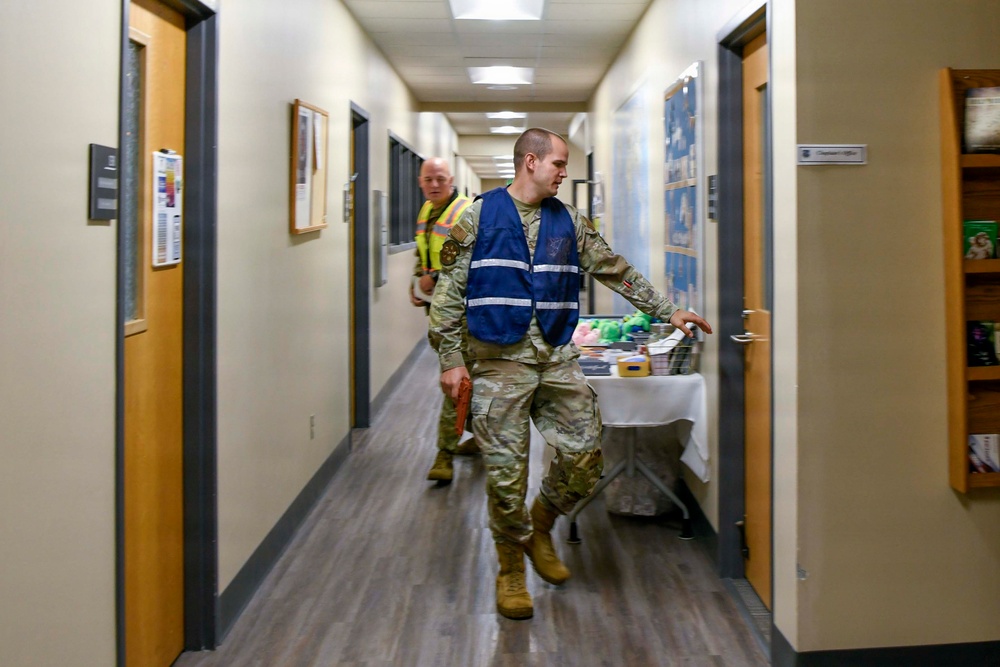 Two individuals in military camouflage uniforms are walking down an indoor hallway of what appears to be a military or government building. The person in the foreground wears a blue reflective safety vest over their uniform, while the person behind wears a yellow high-visibility safety vest. A folding table with various items is visible along the right side of the corridor.