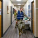Two individuals in military camouflage uniforms are walking down an indoor hallway of what appears to be a military or government building. The person in the foreground wears a blue reflective safety vest over their uniform, while the person behind wears a yellow high-visibility safety vest. A folding table with various items is visible along the right side of the corridor.