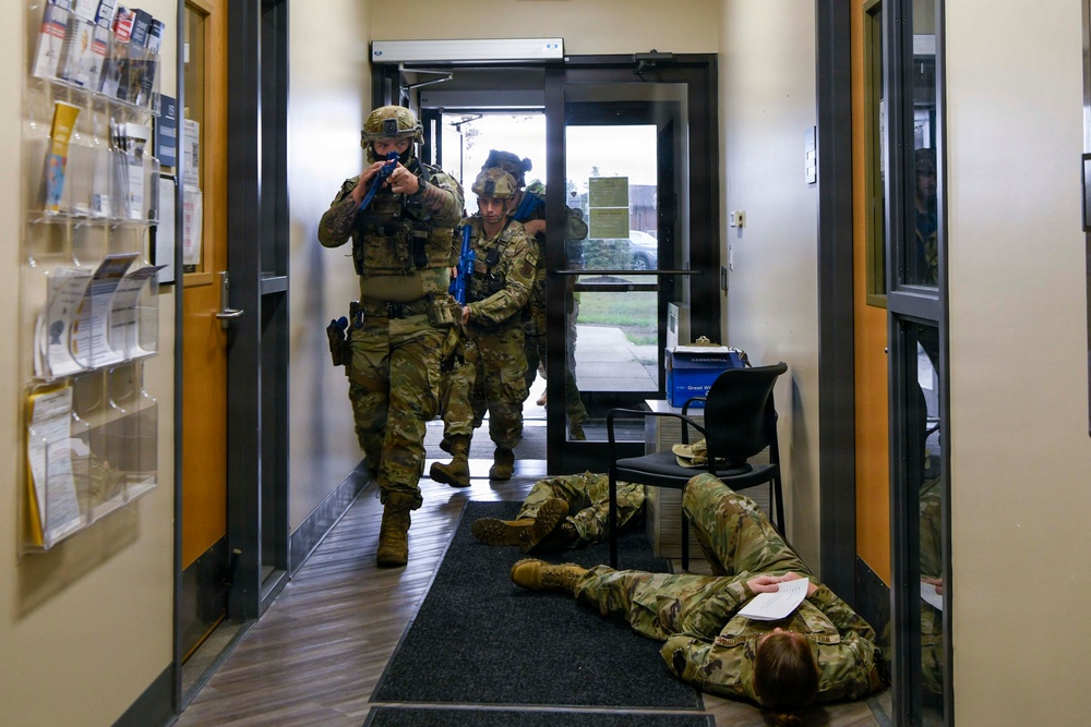 The image depicts a military or law enforcement training exercise taking place inside what appears to be an office or administrative building. Two armed personnel in full tactical gear and camouflage uniforms are moving through a hallway while a third person in matching uniform lies on the floor, simulating a casualty. The blue-colored training weapons and controlled environment confirm this is a drill rather than an actual incident.