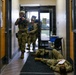 The image depicts a military or law enforcement training exercise taking place inside what appears to be an office or administrative building. Two armed personnel in full tactical gear and camouflage uniforms are moving through a hallway while a third person in matching uniform lies on the floor, simulating a casualty. The blue-colored training weapons and controlled environment confirm this is a drill rather than an actual incident.