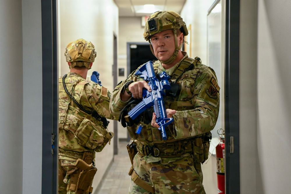 Two military personnel in OCP (Operational Camouflage Pattern) uniforms are conducting what appears to be a building clearing or active shooter response training exercise inside a corridor. The foreground soldier is holding a blue training/inert replica firearm, which is a standard indicator of a simulated training environment rather than a live operation. Both personnel are fully equipped with tactical gear, helmets, and body armor.