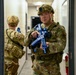 Two military personnel in OCP (Operational Camouflage Pattern) uniforms are conducting what appears to be a building clearing or active shooter response training exercise inside a corridor. The foreground soldier is holding a blue training/inert replica firearm, which is a standard indicator of a simulated training environment rather than a live operation. Both personnel are fully equipped with tactical gear, helmets, and body armor.