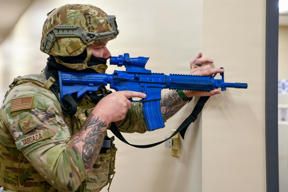 A U.S. Air Force staff sergeant named Taboada is conducting a tactical training exercise indoors, holding a blue training rifle (inert/simunition weapon used for drills). The soldier is in full multicam combat gear including a tactical helmet, face covering, and plate carrier, and is positioned against a wall in a shooting stance. The blue-colored weapon indicates this is a non-lethal training aid used in force-on-force or active shooter response exercises.