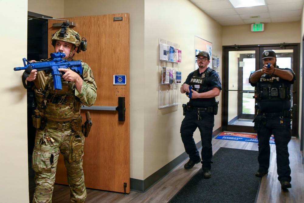 The image depicts what appears to be a law enforcement training exercise taking place inside an office or institutional building. Three personnel are visible: one in full military/tactical camouflage gear holding a blue training rifle, and two officers in black police uniforms holding blue training handguns. The blue-colored weapons are a standard indicator of simulated training firearms (inert or Airsoft/Simunition), confirming this is a drill rather than a live operation.