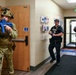 The image depicts what appears to be a law enforcement training exercise taking place inside an office or institutional building. Three personnel are visible: one in full military/tactical camouflage gear holding a blue training rifle, and two officers in black police uniforms holding blue training handguns. The blue-colored weapons are a standard indicator of simulated training firearms (inert or Airsoft/Simunition), confirming this is a drill rather than a live operation.