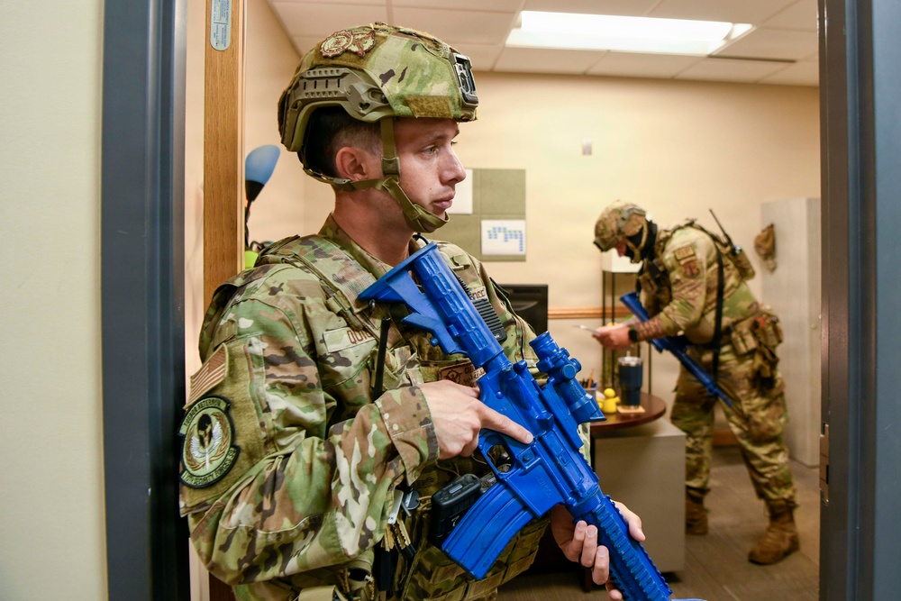 Two military or law enforcement personnel in full MultiCam/OCP camouflage uniforms and tactical helmets are conducting what appears to be a training exercise inside an office or institutional building. Both individuals are carrying bright blue training/simulation rifles, which are non-lethal replica weapons used exclusively for drills. The setting appears to be a room-clearing or active-shooter response training scenario.