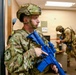 Two military or law enforcement personnel in full MultiCam/OCP camouflage uniforms and tactical helmets are conducting what appears to be a training exercise inside an office or institutional building. Both individuals are carrying bright blue training/simulation rifles, which are non-lethal replica weapons used exclusively for drills. The setting appears to be a room-clearing or active-shooter response training scenario.