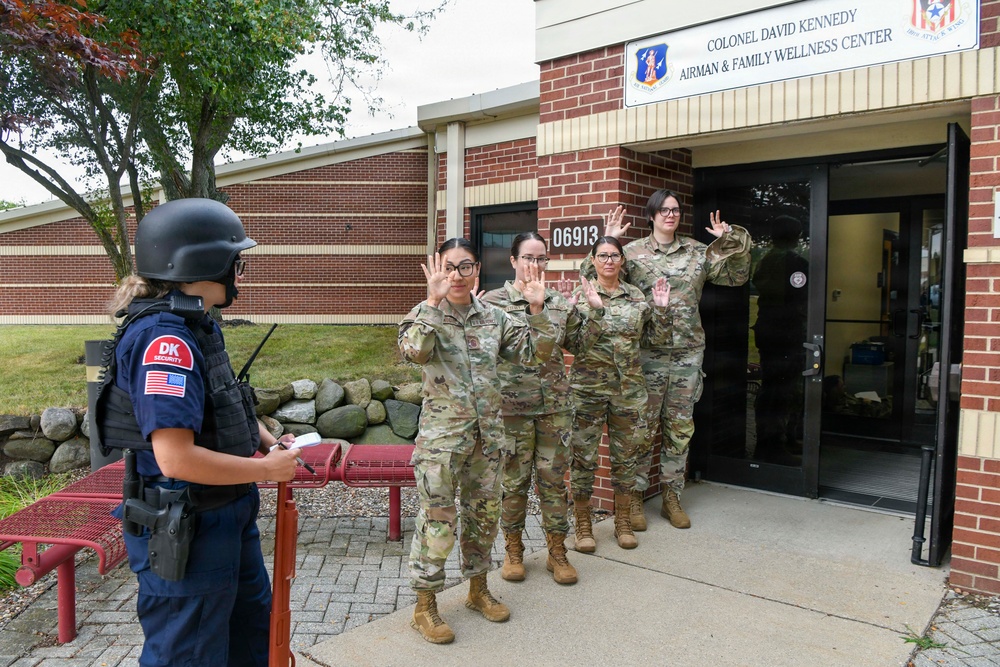A security drill or active shooter exercise is taking place outside the Colonel David Kennedy Airman &amp; Family Wellness Center (Building 06913) on a military installation. A DK Security officer wearing tactical gear and a ballistic helmet is facing four military personnel in OCP (Operational Camouflage Pattern) uniforms who have their hands raised. This appears to be a training scenario simulating an emergency response or evacuation procedure.