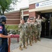 A security drill or active shooter exercise is taking place outside the Colonel David Kennedy Airman &amp; Family Wellness Center (Building 06913) on a military installation. A DK Security officer wearing tactical gear and a ballistic helmet is facing four military personnel in OCP (Operational Camouflage Pattern) uniforms who have their hands raised. This appears to be a training scenario simulating an emergency response or evacuation procedure.