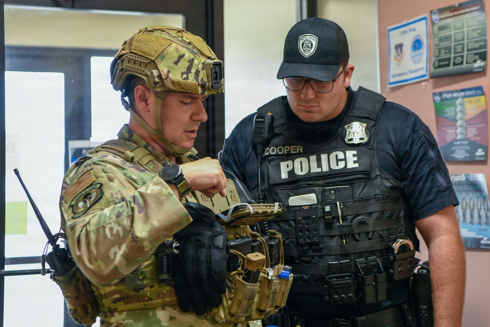 Two law enforcement/security personnel are shown in an indoor setting, appearing to review a handheld device or firearm. One individual is wearing full military tactical gear including a multicam helmet and camouflage uniform, while the other is a police officer identified by name tag 'Cooper' wearing a tactical vest. They appear to be engaged in a briefing, equipment check, or coordination activity.