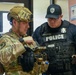 Two law enforcement/security personnel are shown in an indoor setting, appearing to review a handheld device or firearm. One individual is wearing full military tactical gear including a multicam helmet and camouflage uniform, while the other is a police officer identified by name tag 'Cooper' wearing a tactical vest. They appear to be engaged in a briefing, equipment check, or coordination activity.