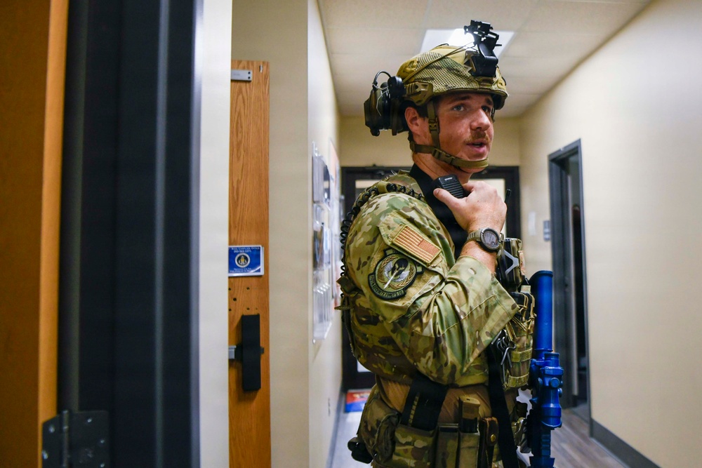 A military or law enforcement operator in full tactical gear is positioned in an indoor hallway, appearing to communicate via radio. The individual is wearing OCP (Operational Camouflage Pattern) uniform with patches, a tactical helmet with mounted light, and a plate carrier vest. The setting appears to be a training exercise or active operation inside a building.