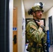 A military or law enforcement operator in full tactical gear is positioned in an indoor hallway, appearing to communicate via radio. The individual is wearing OCP (Operational Camouflage Pattern) uniform with patches, a tactical helmet with mounted light, and a plate carrier vest. The setting appears to be a training exercise or active operation inside a building.