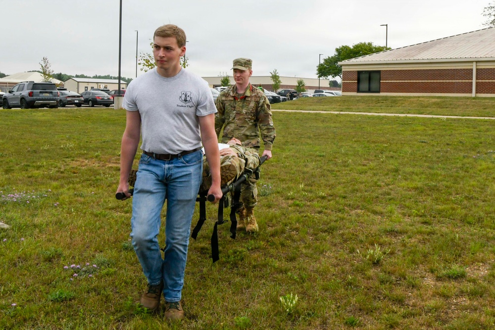 Two individuals are carrying a military litter (stretcher) across a grassy field near a brick building and parking lot. The person in front is wearing civilian clothes with a gray 'Student Flight' t-shirt and jeans, while the person in the rear is wearing full military camouflage (OCP/MultiCam) uniform. This appears to be a training exercise, likely involving casualty evacuation or combat lifesaver skills.