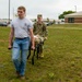 Two individuals are carrying a military litter (stretcher) across a grassy field near a brick building and parking lot. The person in front is wearing civilian clothes with a gray 'Student Flight' t-shirt and jeans, while the person in the rear is wearing full military camouflage (OCP/MultiCam) uniform. This appears to be a training exercise, likely involving casualty evacuation or combat lifesaver skills.