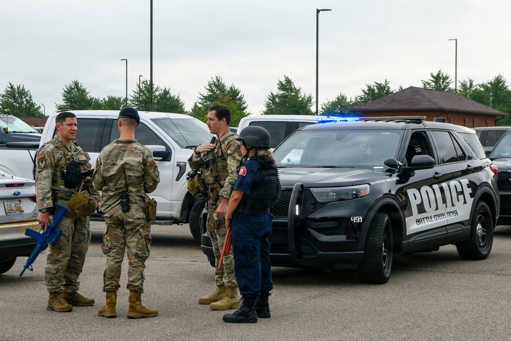 The image shows a group of military personnel in camouflage uniforms and one individual in dark tactical gear standing in a parking lot near a Battle Creek Area Police vehicle with active blue lights. The personnel appear to be engaged in a briefing or coordination discussion, with some holding training/blue-barrel firearms. This appears to be a joint law enforcement and military training exercise or coordinated response scenario.
