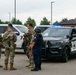 The image shows a group of military personnel in camouflage uniforms and one individual in dark tactical gear standing in a parking lot near a Battle Creek Area Police vehicle with active blue lights. The personnel appear to be engaged in a briefing or coordination discussion, with some holding training/blue-barrel firearms. This appears to be a joint law enforcement and military training exercise or coordinated response scenario.