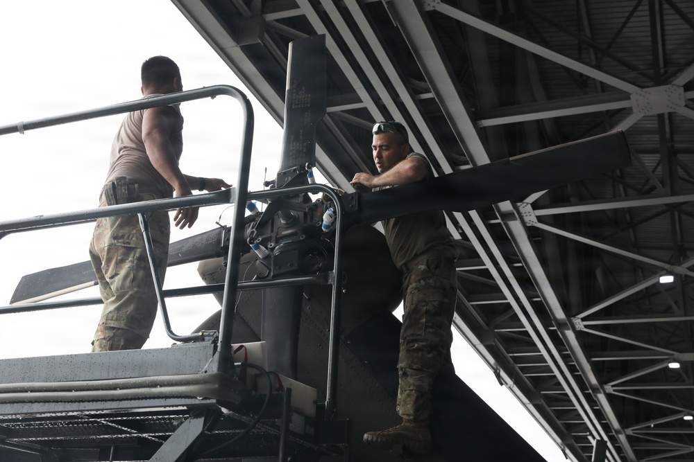 Two military personnel in camouflage uniforms are working on what appears to be a large piece of military equipment or machinery inside a hangar or covered structure. Both individuals are wearing multicam/OCP pattern uniforms and are positioned at an elevated platform. They appear to be performing maintenance or assembly tasks on the equipment.
