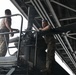Two military personnel in camouflage uniforms are working on what appears to be a large piece of military equipment or machinery inside a hangar or covered structure. Both individuals are wearing multicam/OCP pattern uniforms and are positioned at an elevated platform. They appear to be performing maintenance or assembly tasks on the equipment.